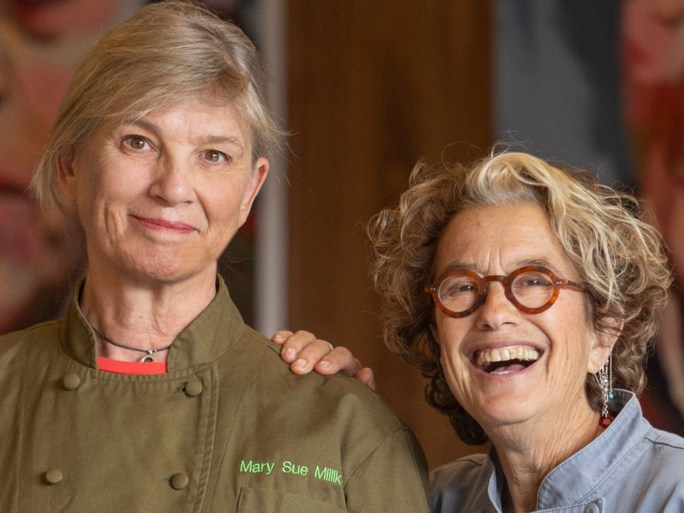 Chef Mary Sue Milliken in an olive green embroidered chef's coat standing alongside Chef Susan Feniger, who wears round tortoiseshell glasses and a grey chef's coat, both smiling warmly.
