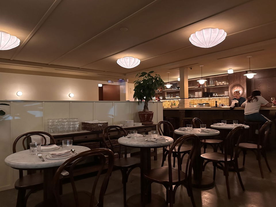 A warmly lit restaurant dining room with marble-topped round tables, bentwood chairs, pendant globe lights, and a bar area.