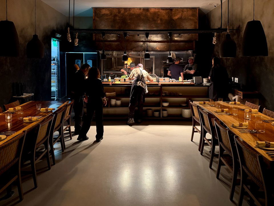 An open-kitchen restaurant dining room with warm wood communal tables, pendant lighting, and chefs working at a live-fire station.