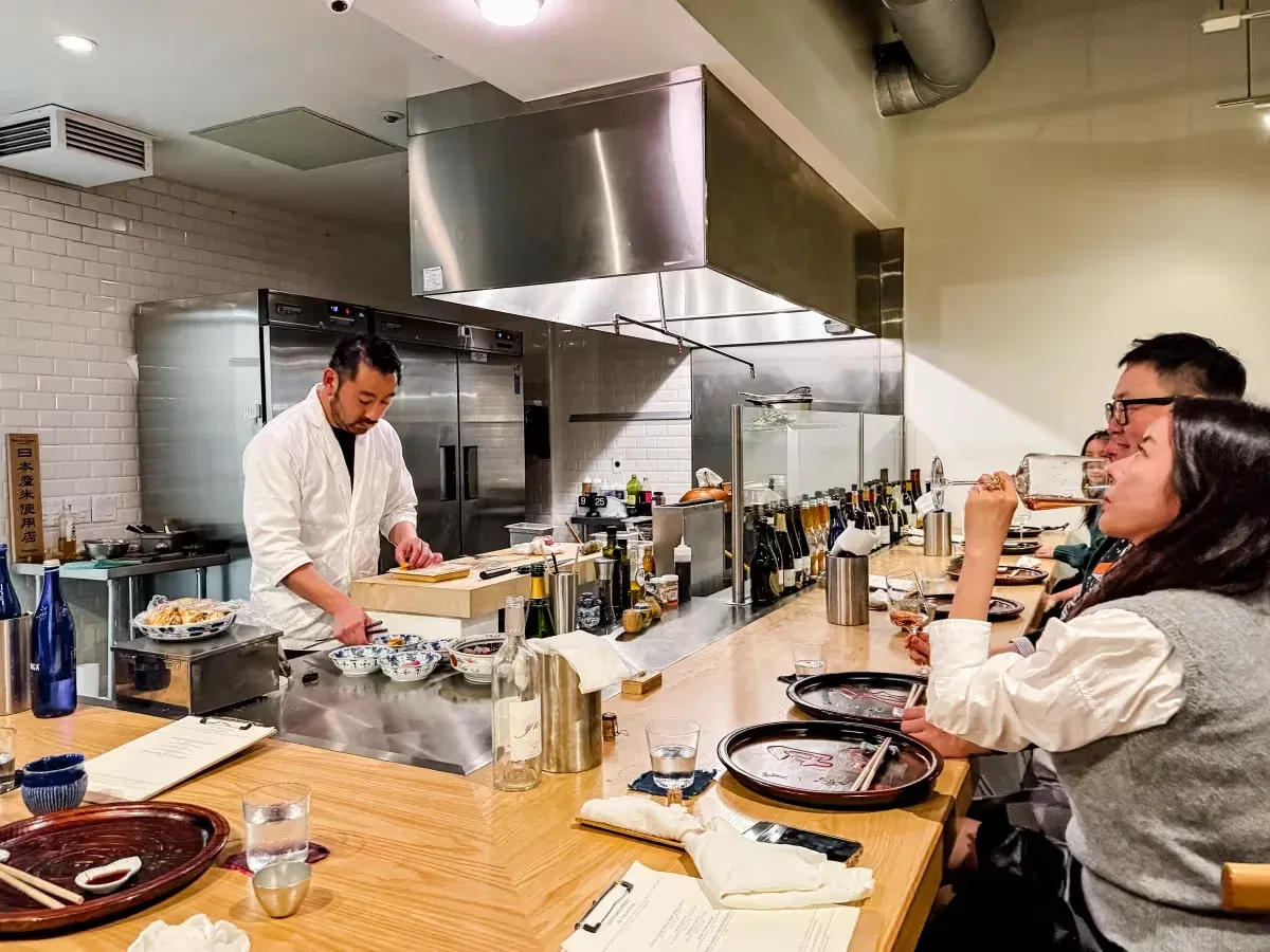 A chef in a white coat prepares food behind a counter, with customers seated at the counter, one drinking from a glass.