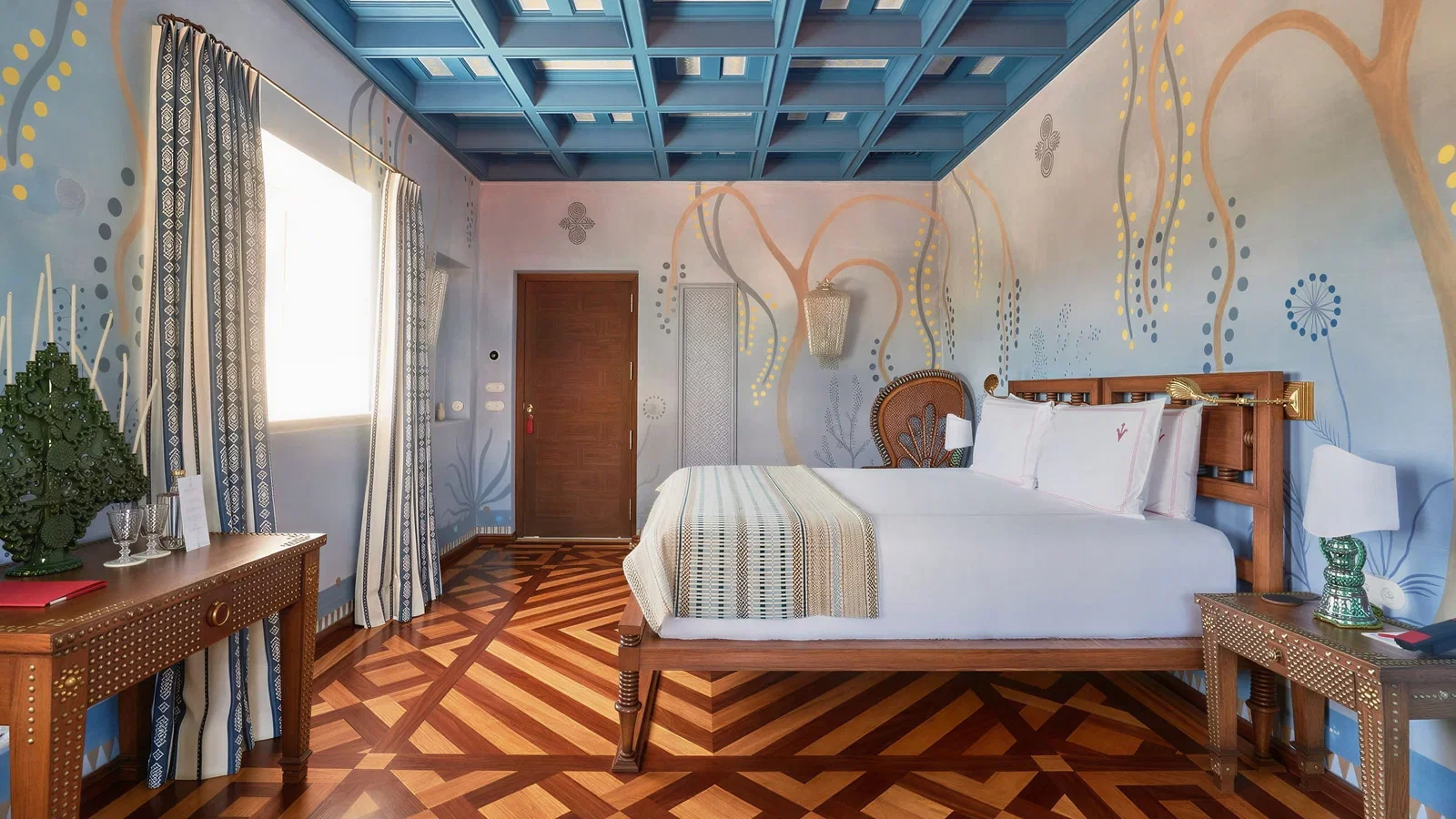 A bedroom at Vermelho Melides features a wooden bed, patterned curtains, and a blue coffered ceiling, with a desk and ornate decor.