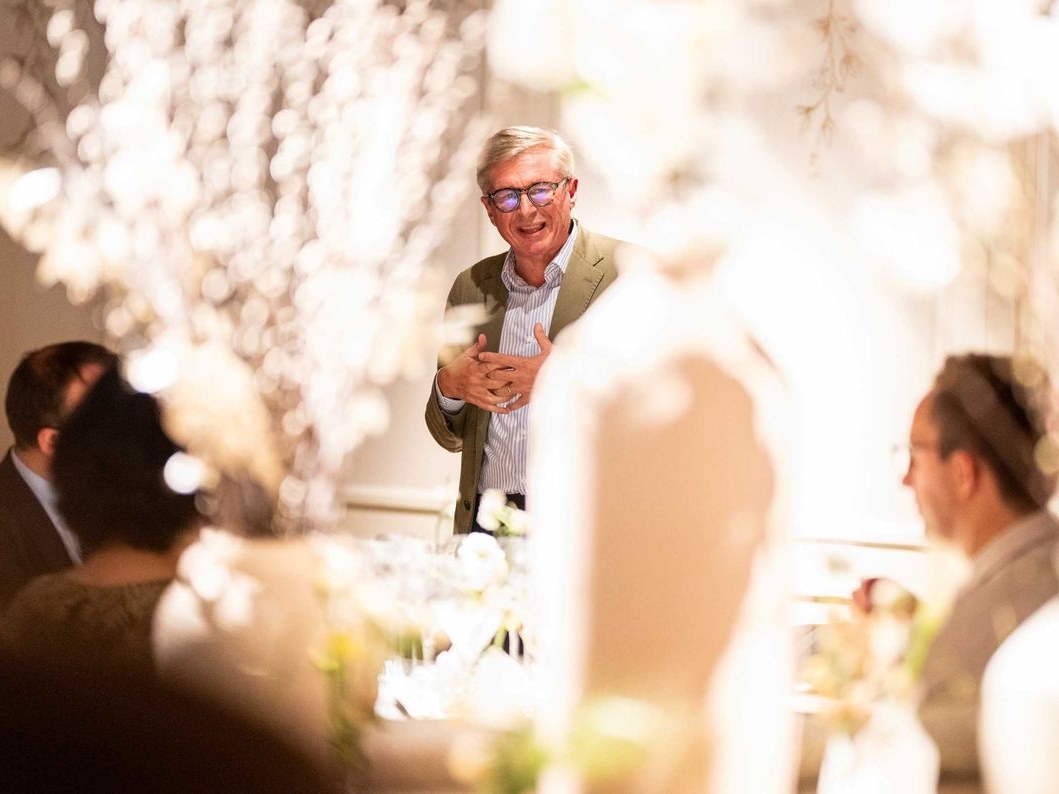 An older man with white hair and glasses wearing a tan blazer and striped shirt is speaking at an elegant event with white floral decorations and seat