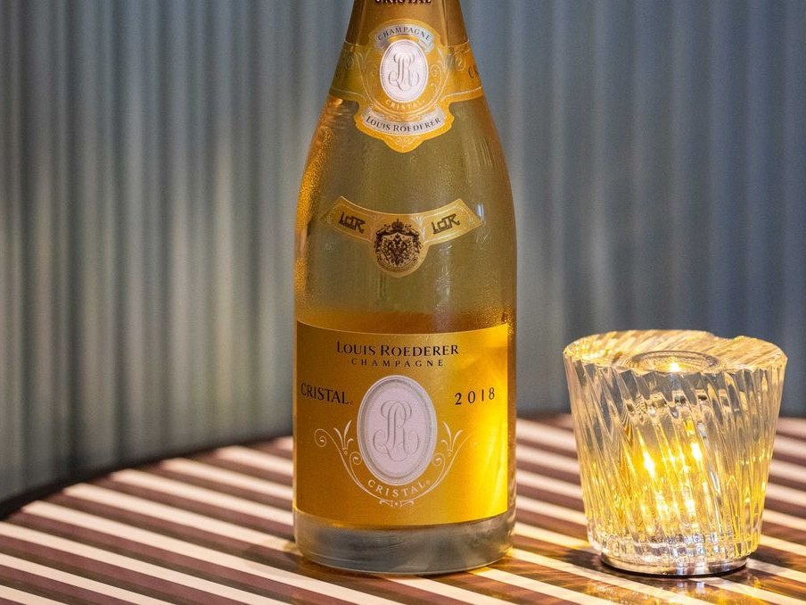 A bottle of Louis Roederer Cristal 2018 champagne with a crystal glass on a striped surface, matching the article subject via visible label text and s