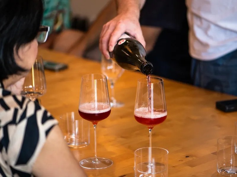 A hand pouring red/rosé wine from a dark bottle into stemmed wine glasses on a wooden communal table, with a patron seated nearby, consistent.