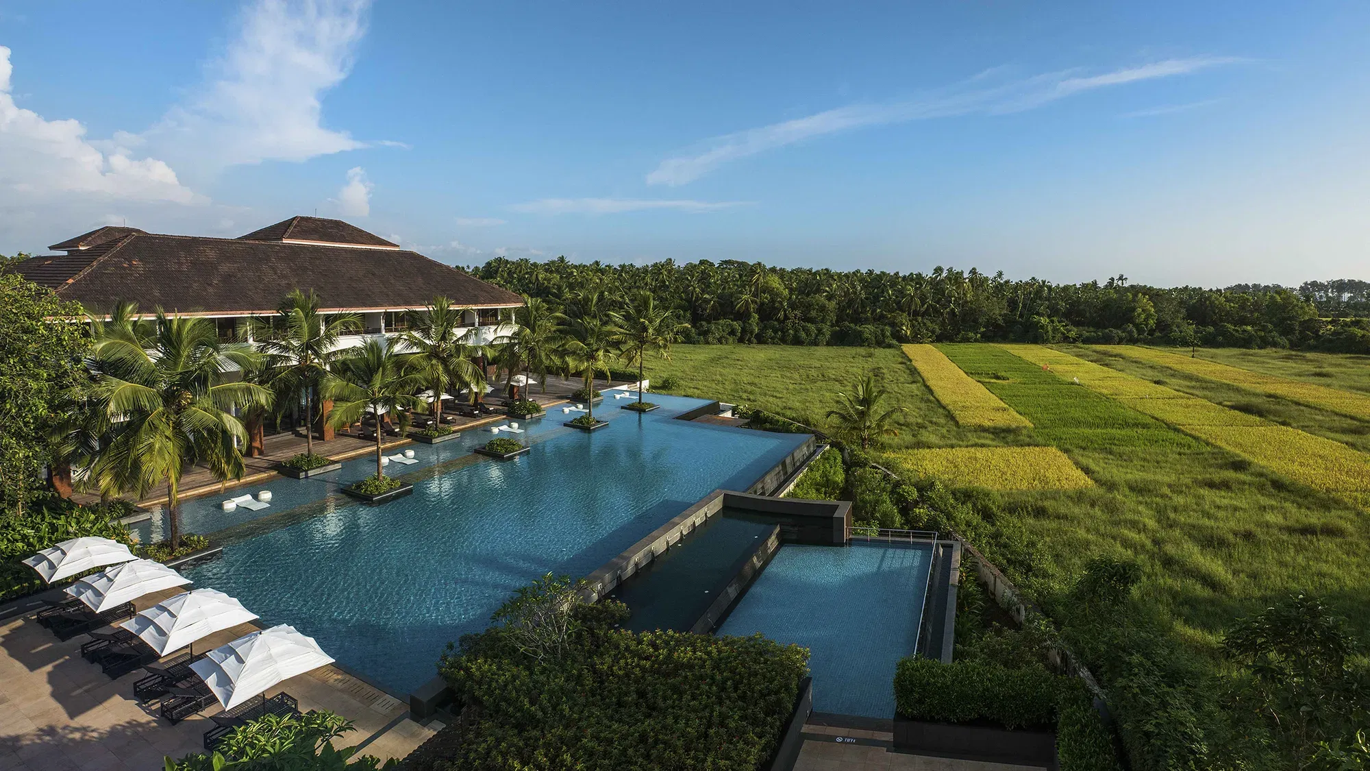 An aerial view of a luxury hotel with a large swimming pool, surrounded by palm trees and lush green fields under a blue sky.