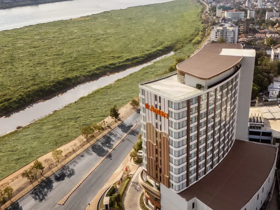 An aerial view of the Amari hotel, a modern high-rise building with a curved facade, situated next to a river and a city.
