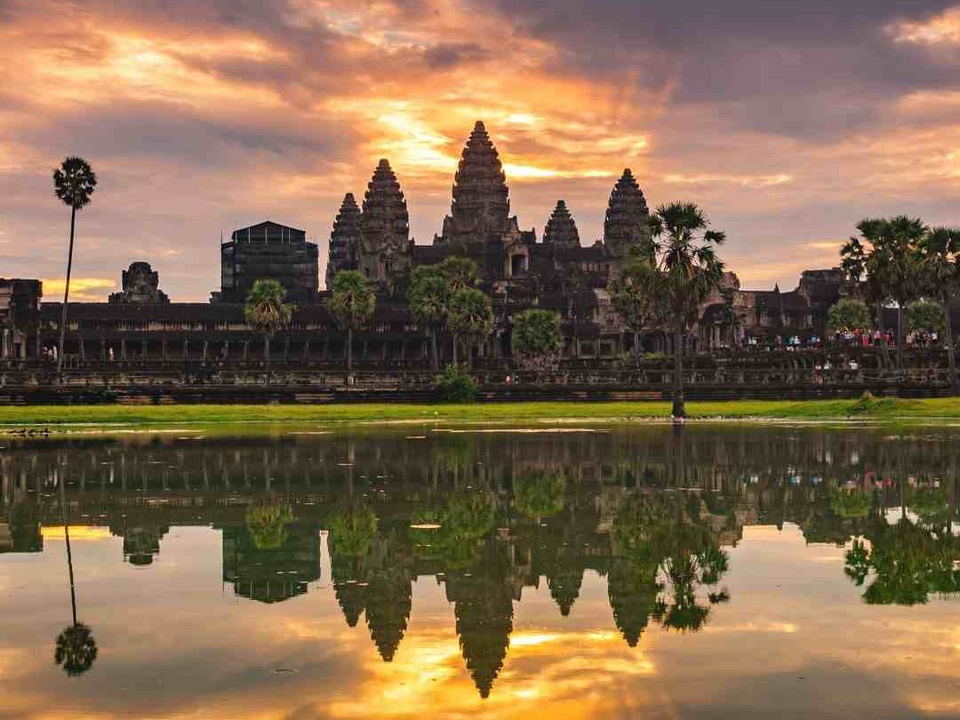 A wide landscape shot of Angkor Wat temple complex at sunrise, reflected in the foreground reservoir, with dramatic orange and purple clouds.