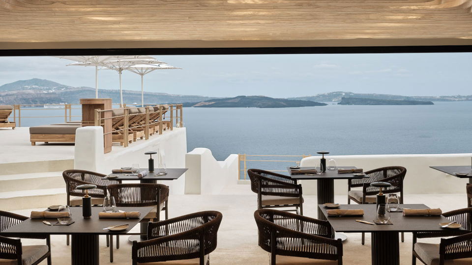 An upscale restaurant dining terrace with dark woven chairs, set tables, white architectural elements, and a panoramic view of a blue sea with islands