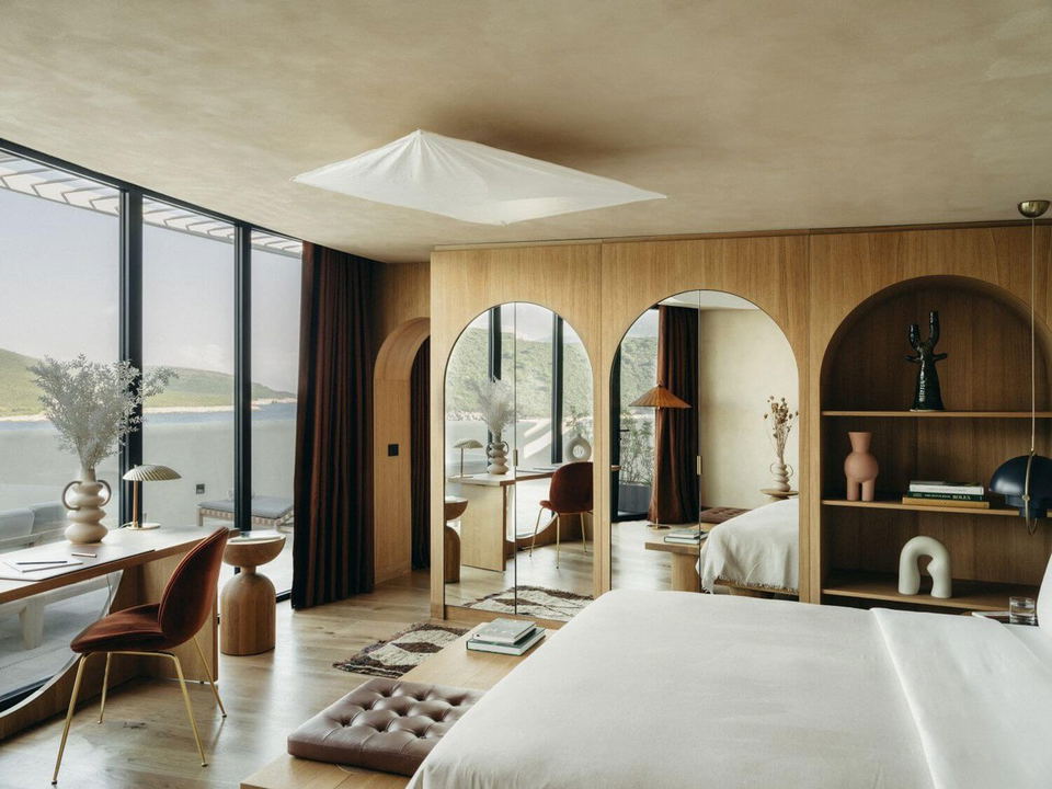 Modern hotel suite with wooden walls, arched doorways, a bed in foreground, seating area, and expansive windows showing water views.