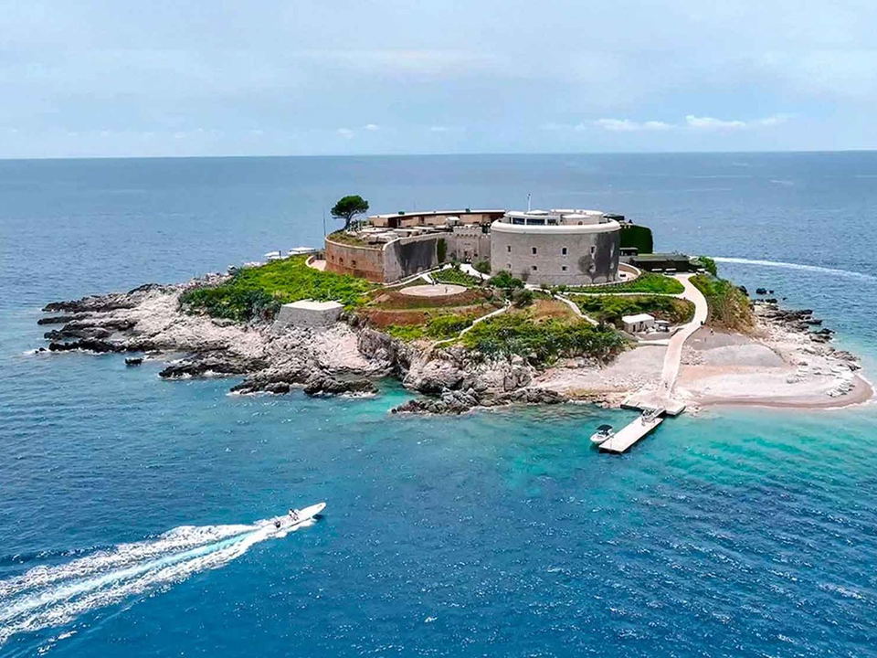 Aerial view of a circular stone fortress on a rocky island in clear blue-green water with boats nearby and a sandy beach area.