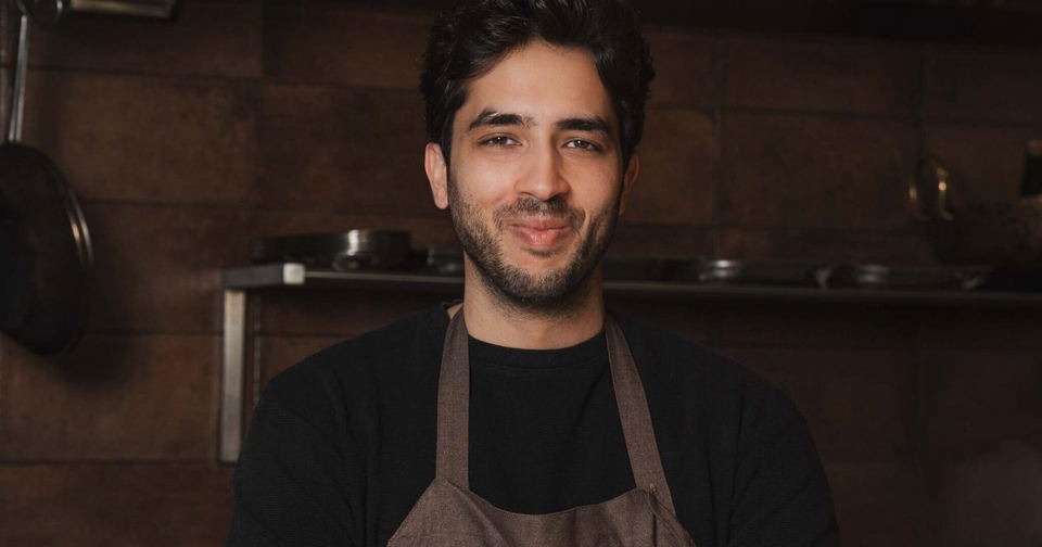 A man with dark hair and beard wearing a black shirt and brown apron in a kitchen with brick walls and shelving.
