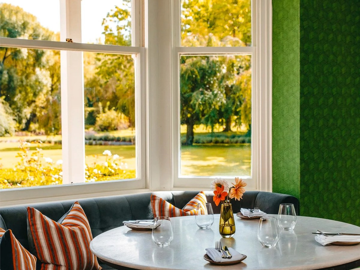 An elegant dining area with a curved banquette, striped cushions, and a marble table set for four, next to large windows overlooking a garden.