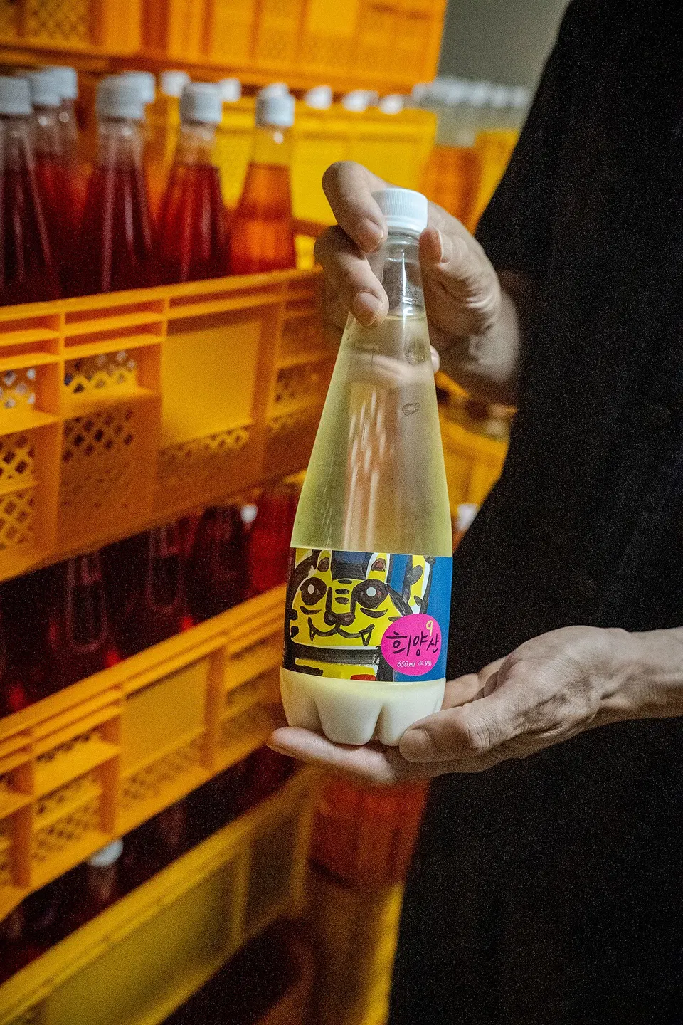 A person holds a bottle of Korean rice wine (makgeolli) with colorful label featuring Korean text, in front of yellow crates containing bottles with r