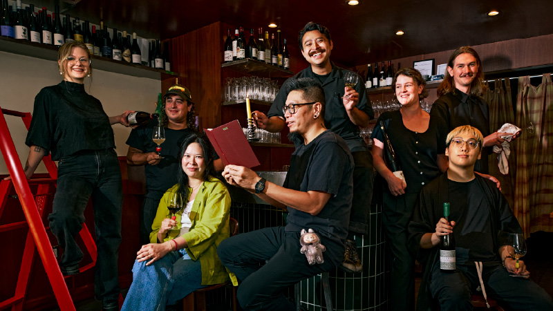 A group of approximately nine hospitality professionals in dark attire posing in a bar/restaurant interior with wine glasses and bottles, published by