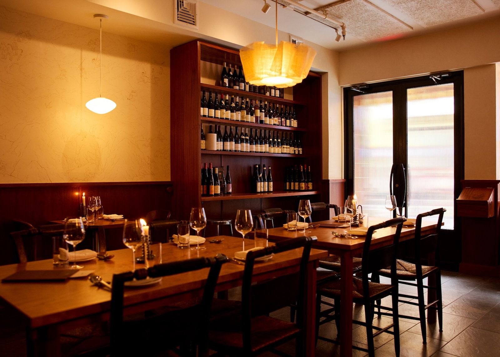 An intimate restaurant dining room with wooden tables set with wine glasses, dark wood wine shelving displaying bottles, warm ambient lighting, and a 