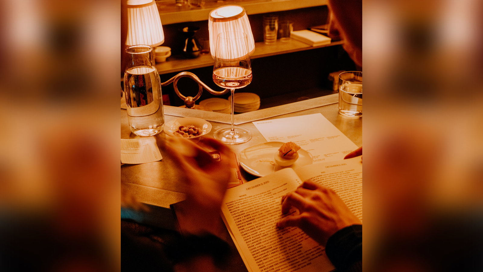 A sommelier or wine professional presenting wine to a guest at an intimate dining table, with the guest reading menu materials