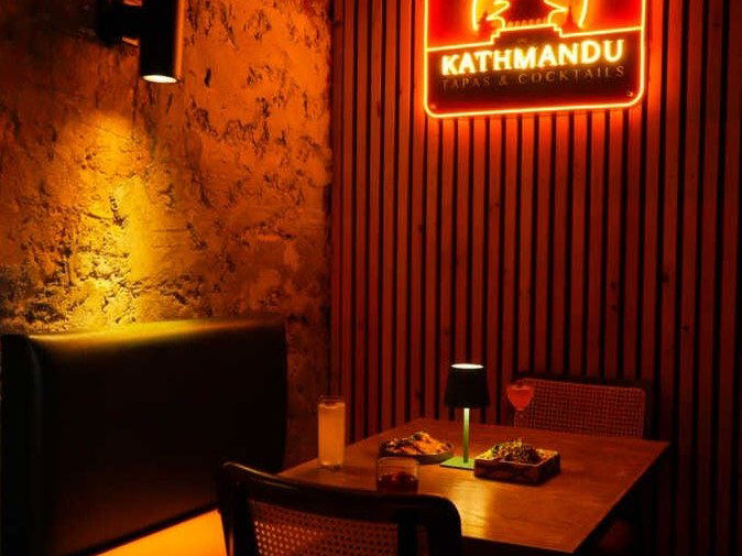 A neon sign for Kathmandu Tapas & Cocktails glows on a slatted wood wall above a table with drinks and food, next to a booth and exposed brick.