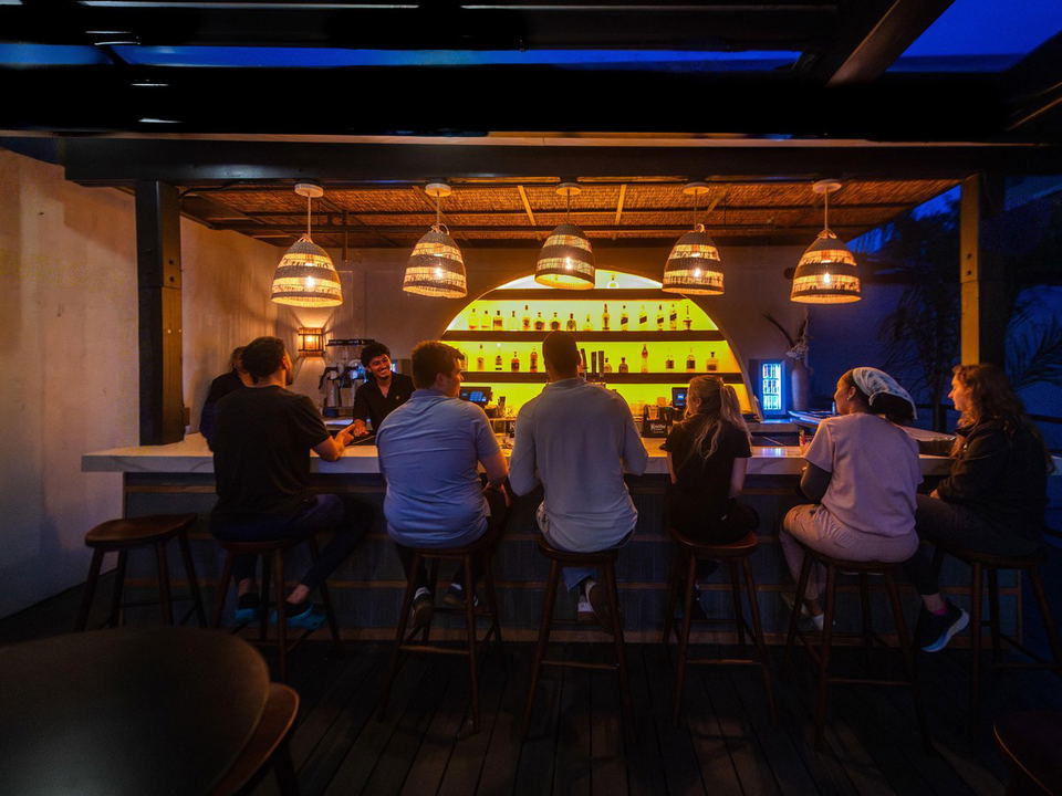 Patrons sit at a warmly lit rooftop bar with glowing yellow liquor shelves and woven pendant lights at twilight.