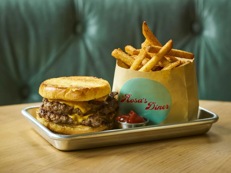 A double smash burger with melted cheese on a toasted bun, served with seasoned fries in a branded paper bag reading "Rosa's Diner" on a metal tray.