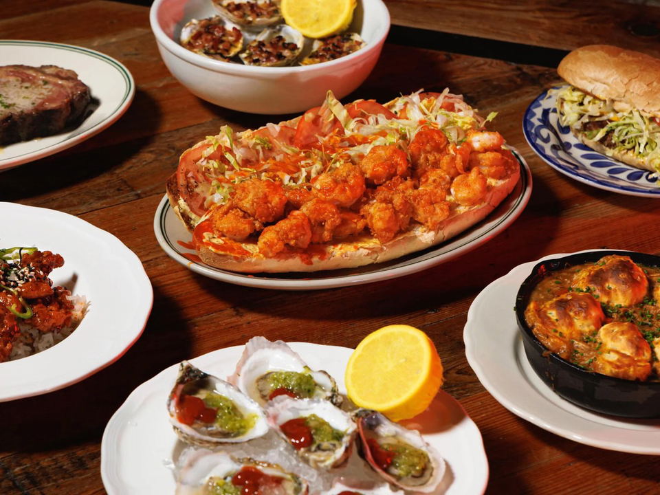 A wooden table laden with various Louisiana-Cajun dishes, including a large shrimp po'boy, raw oysters, a steak, and a skillet.