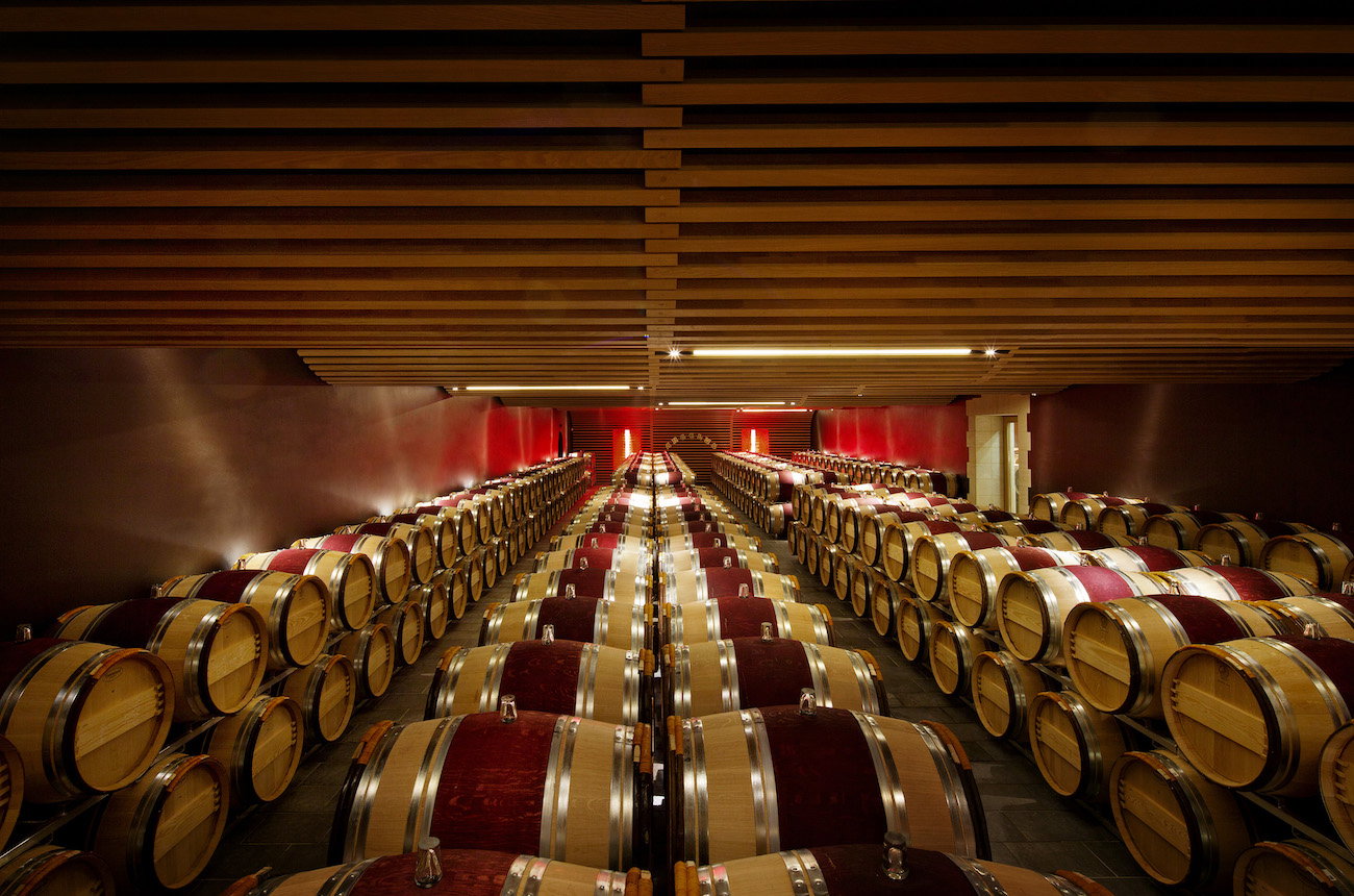 A large barrel cellar with rows of oak wine barrels arranged symmetrically under a dramatic slatted wooden ceiling with linear lighting; the source UR