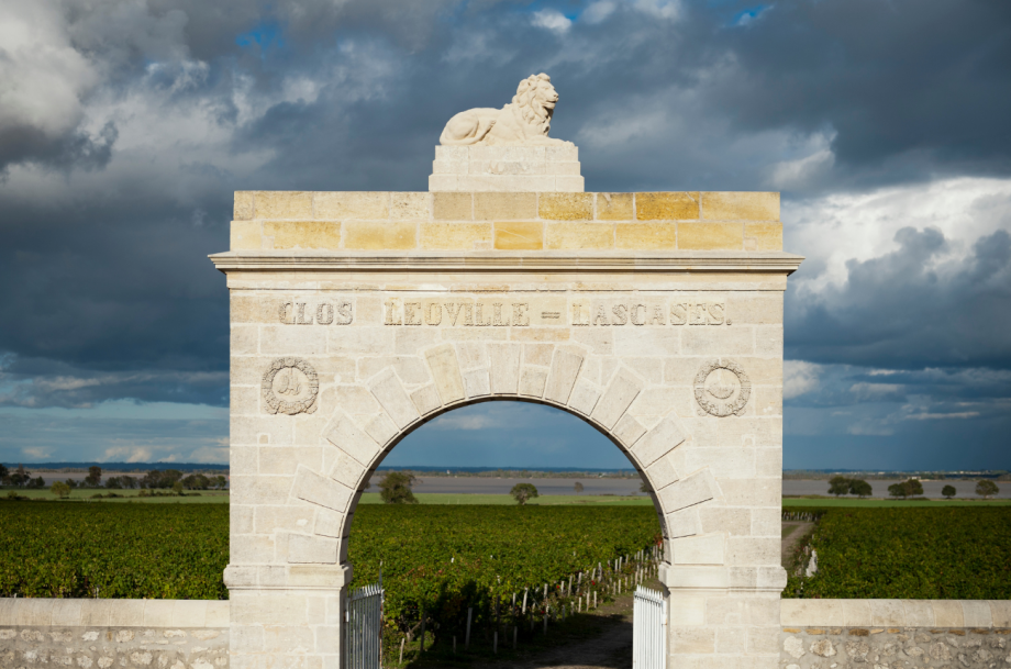Stone archway entrance gate with 'CLOS LEOVILLE - LASCASES' inscription and lion sculpture on top, framing vineyard view beyond; explicit name attesta