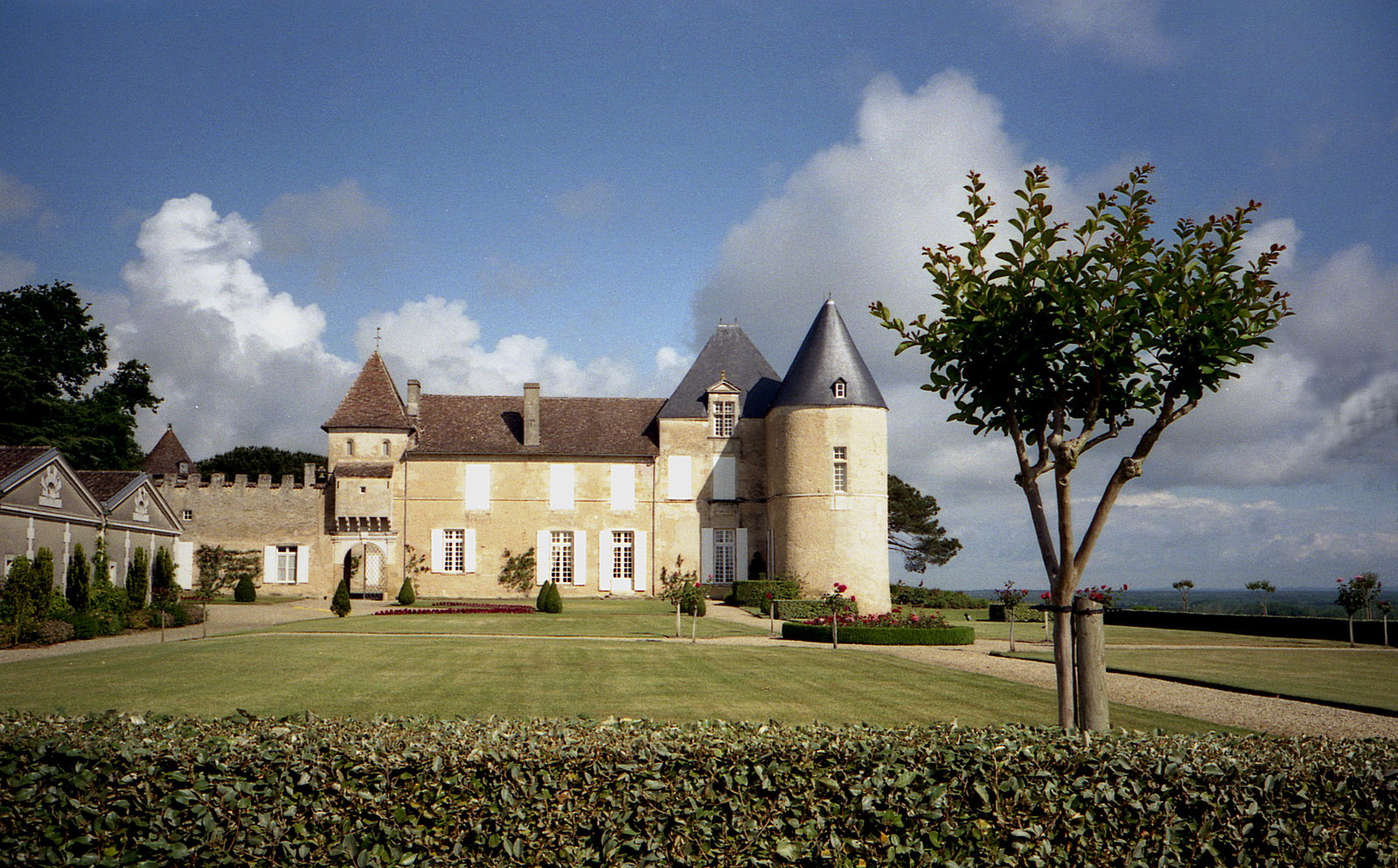 A large château with two round towers topped with pointed roofs, cream-colored stone walls, and manicured lawns, set against a blue sky with white clouds.