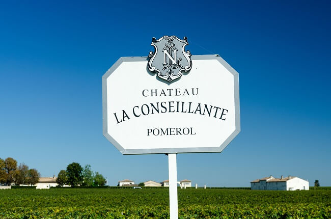 A white estate sign reading 'CHATEAU LA CONSEILLANTE POMEROL' with decorative crest, mounted on a post in front of green vineyard rows under blue sky;