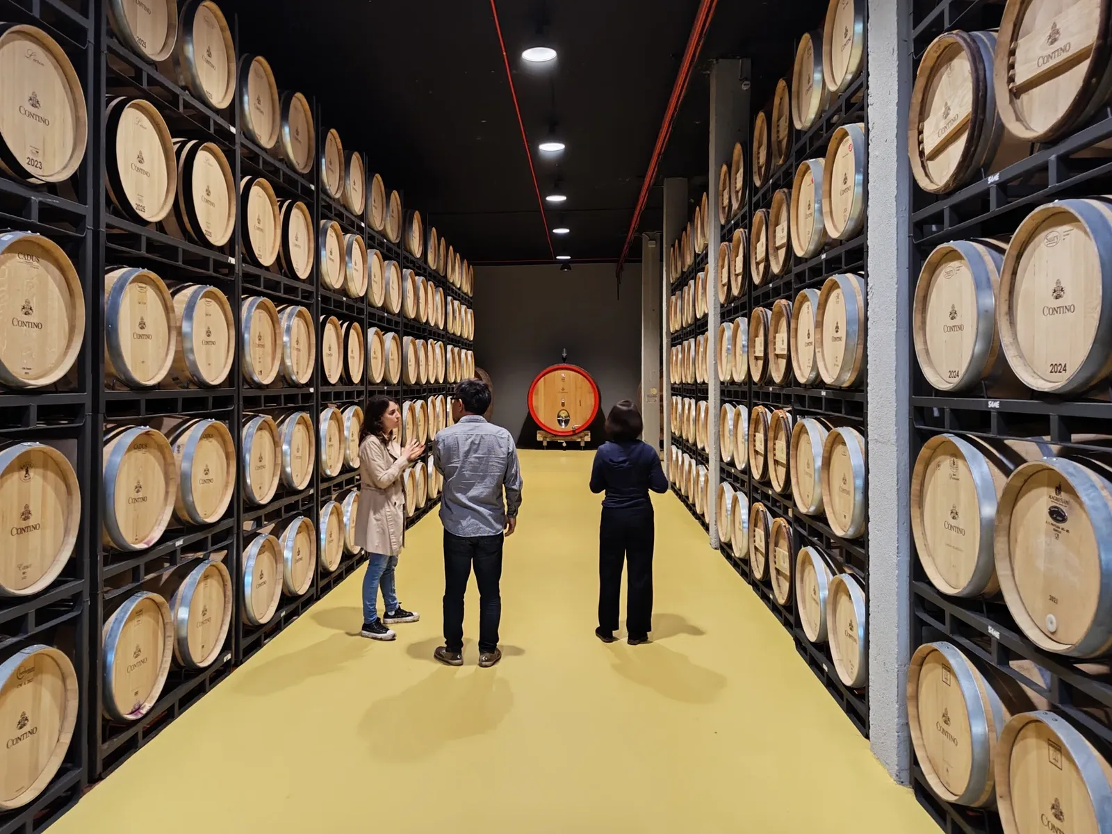 A long, narrow room with rows of wooden wine barrels stacked on metal shelves, and three people standing on a yellow floor.