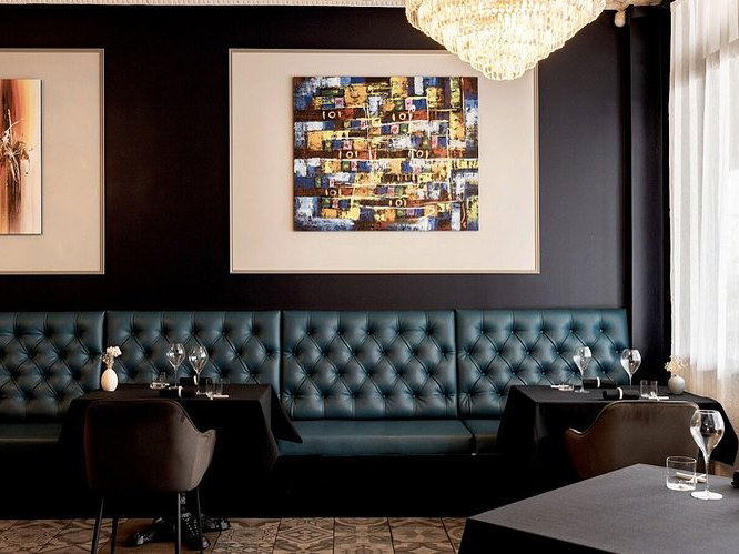 An upscale restaurant dining room with tufted teal leather banquettes, black walls, framed artwork, crystal chandelier, and formally set tables; sourc
