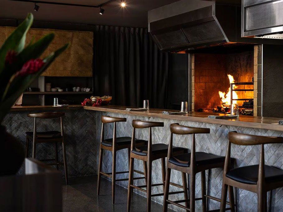 A modern restaurant interior featuring a stone-clad bar counter with wooden bar stools, a dramatic wood-fired oven with visible flames, dark walls, an