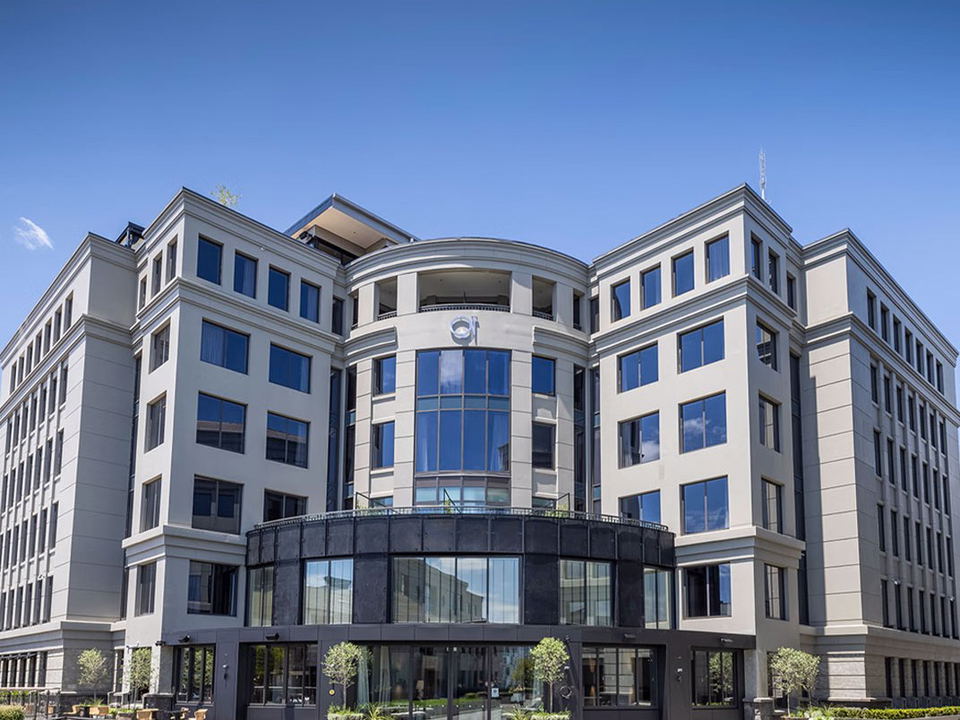 A modern multi-story commercial or hospitality building with curved glass entrance, white facade, and manicured lawn; the source URL explicitly identi