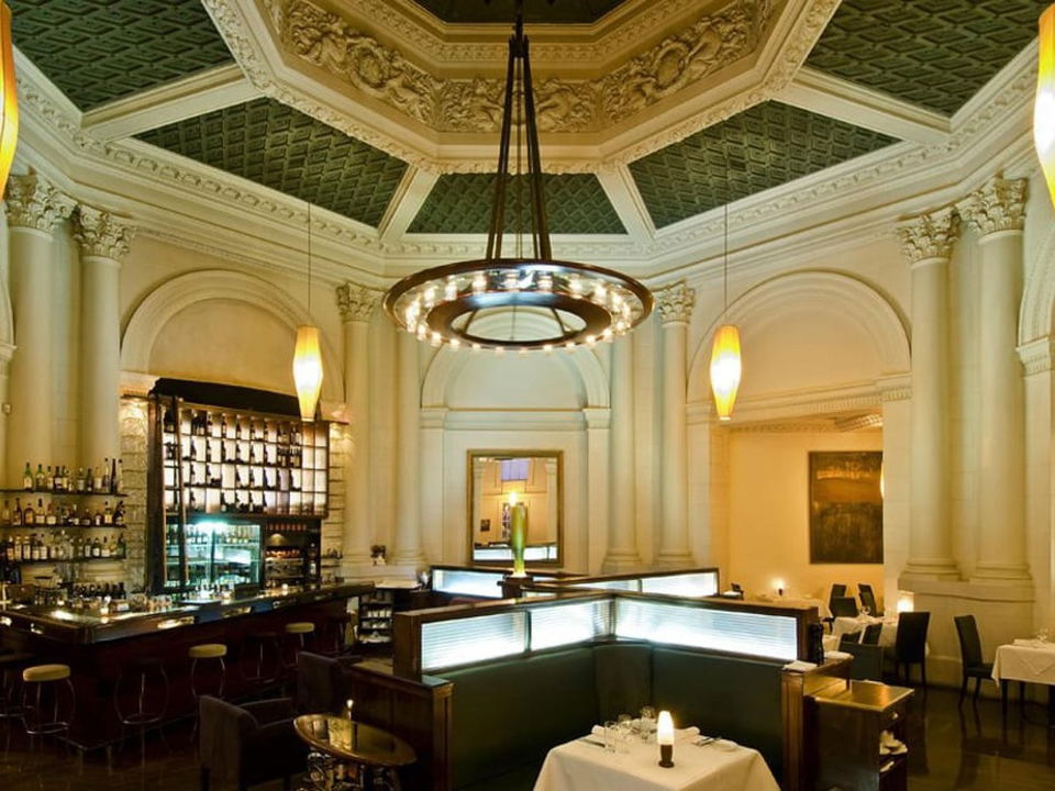 An elegant restaurant dining room with ornate neoclassical architecture, featuring a coffered green ceiling, Corinthian columns, arched alcoves, a cir