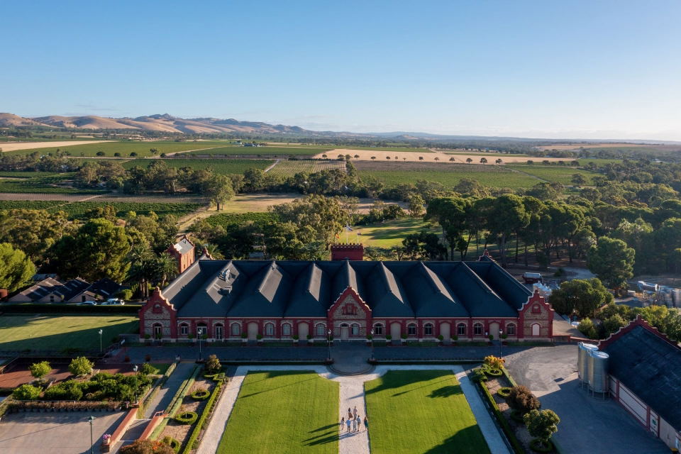 Aerial drone photograph of Chateau Tanunda in the Barossa Valley, South Australia, showing the grand red-brick winery building with a dark Dutch-gabled roof, formal green lawns with a central path, and rows of vineyards extending to rolling hills under a clear blue sky.