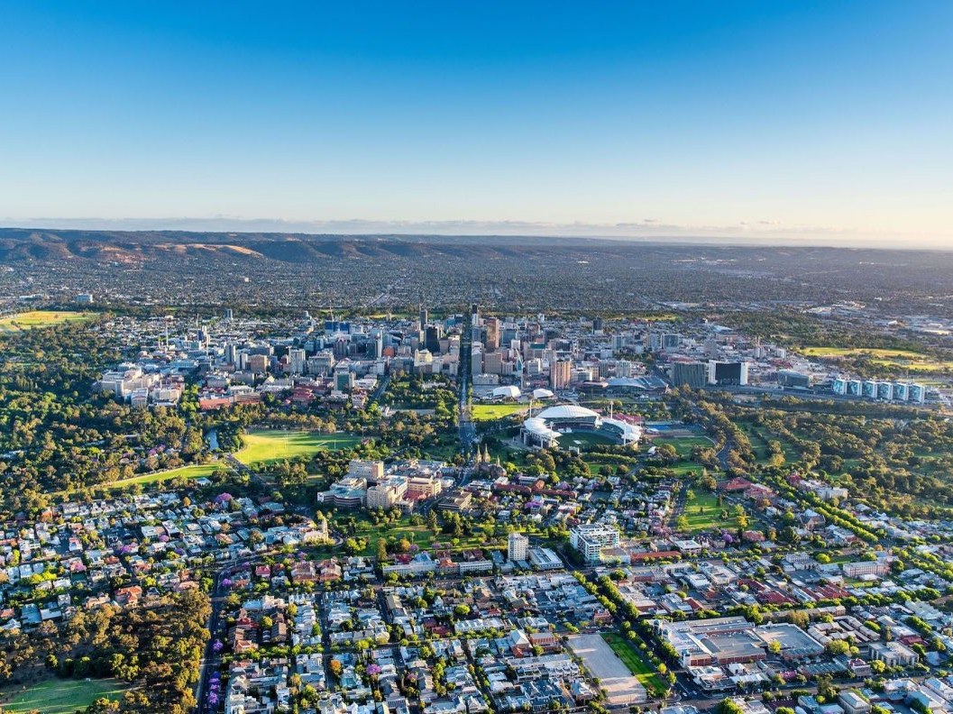 An aerial view of Adelaide, South Australia, with a city skyline, green parks, and residential areas under a clear blue sky.