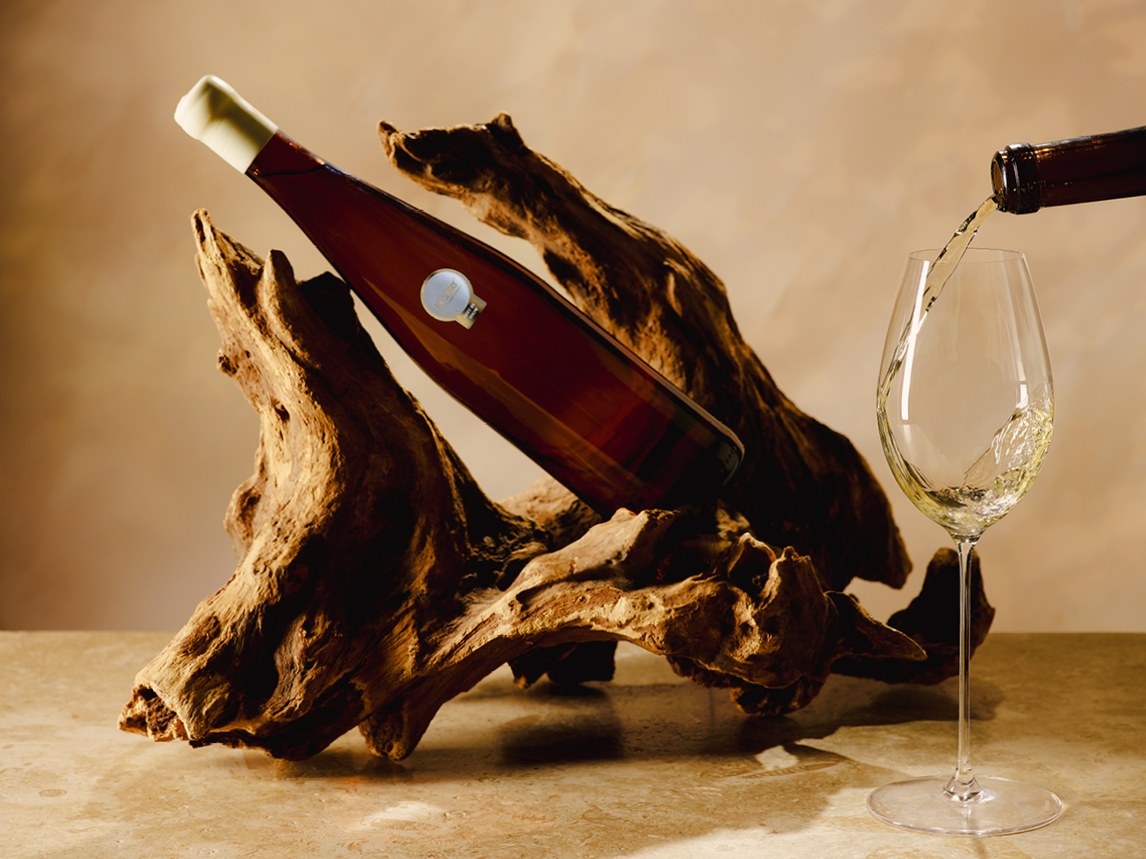 A dark wine bottle sits in a piece of weathered driftwood while white wine is poured into a stemmed glass beside it.