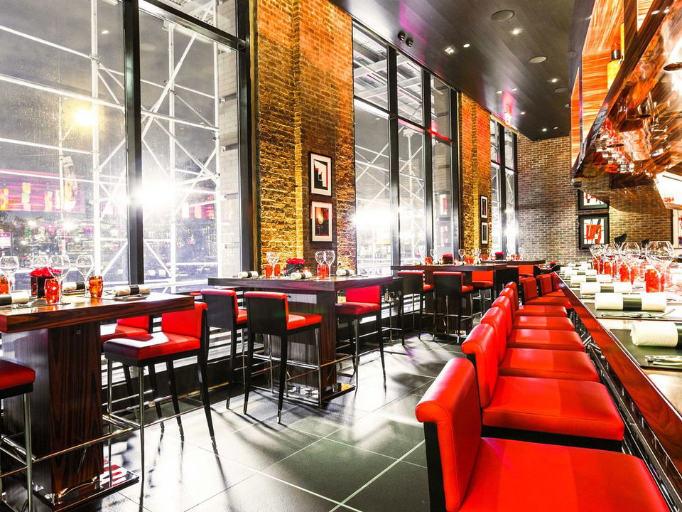Modern restaurant interior with counter seating featuring bright red upholstered seats, open kitchen design with copper/orange metallic surfaces, expo