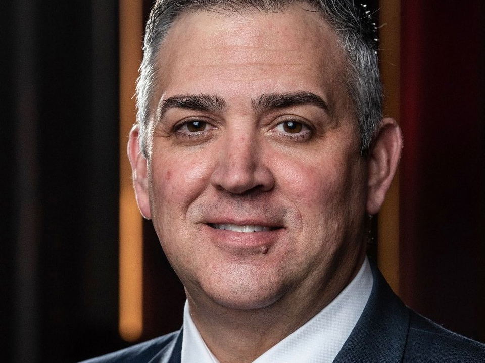 Professional corporate portrait of a man in a dark suit and patterned pink tie against a dark background; source page from Michelin Guide explicitly a