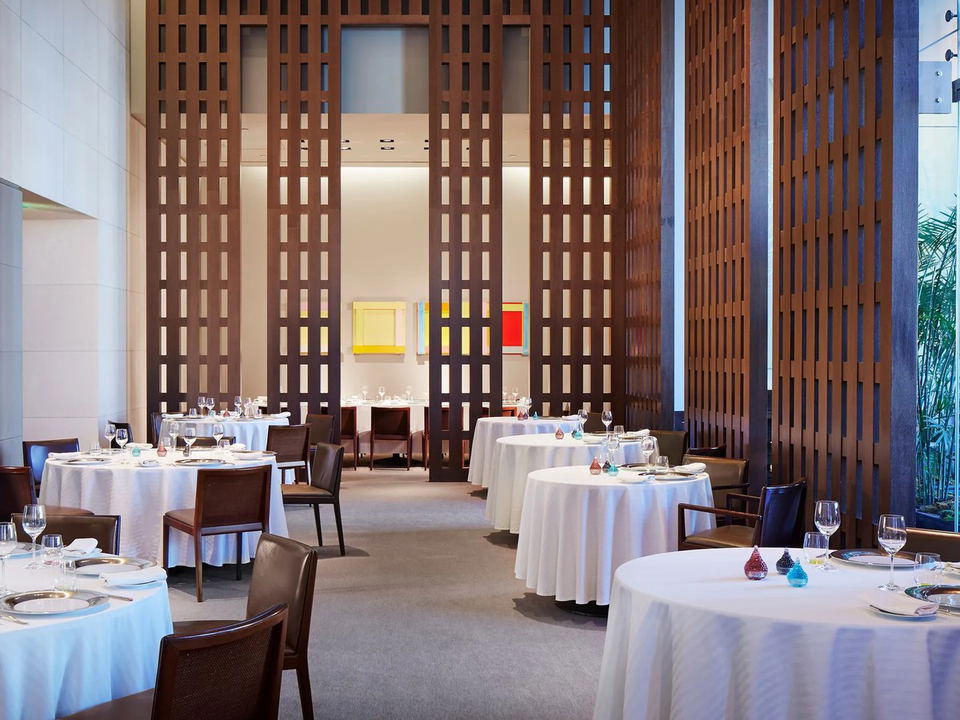 Fine dining restaurant interior with round tables set with white tablecloths, brown leather chairs, tall wooden lattice room dividers, and colorful artwork on walls.