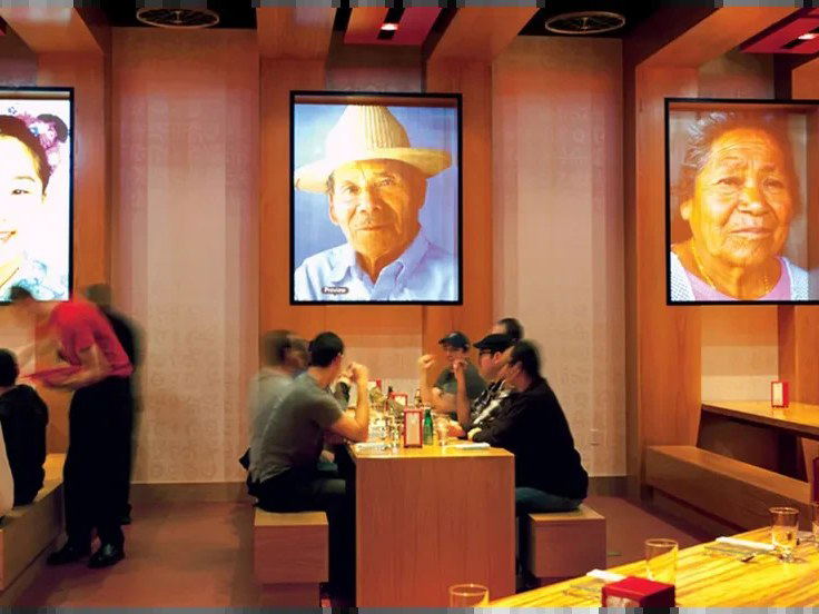 Modern casual dining room with large backlit portrait photographs on walls, communal-style seating with diners at tables; Condé Nast Traveler page exp