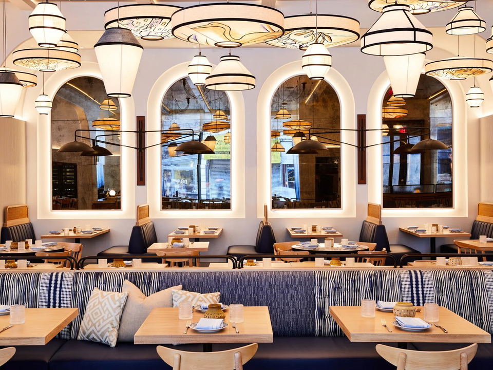 Modern restaurant dining room with distinctive tiered pendant lighting, arched windows, banquette seating with striped upholstery, and set tables; sou