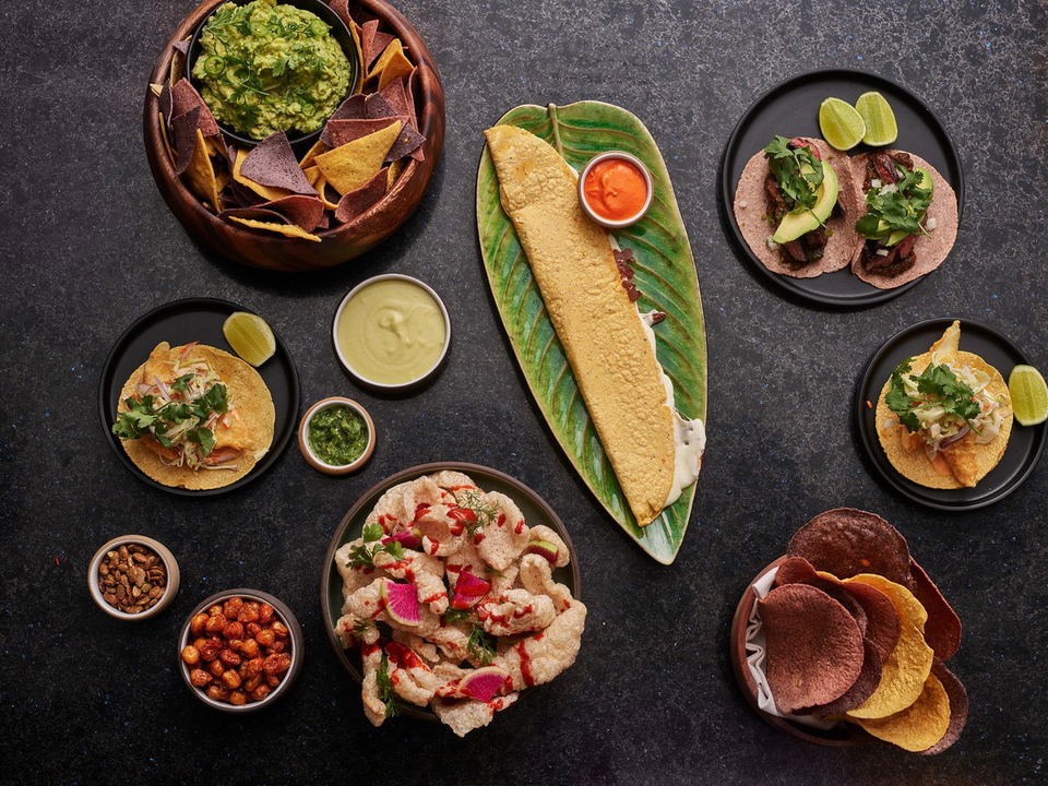 A flat-lay composition of Mexican cuisine including tacos, guacamole with chips, chicharrones, and various sauces on a dark surface; trusted editorial