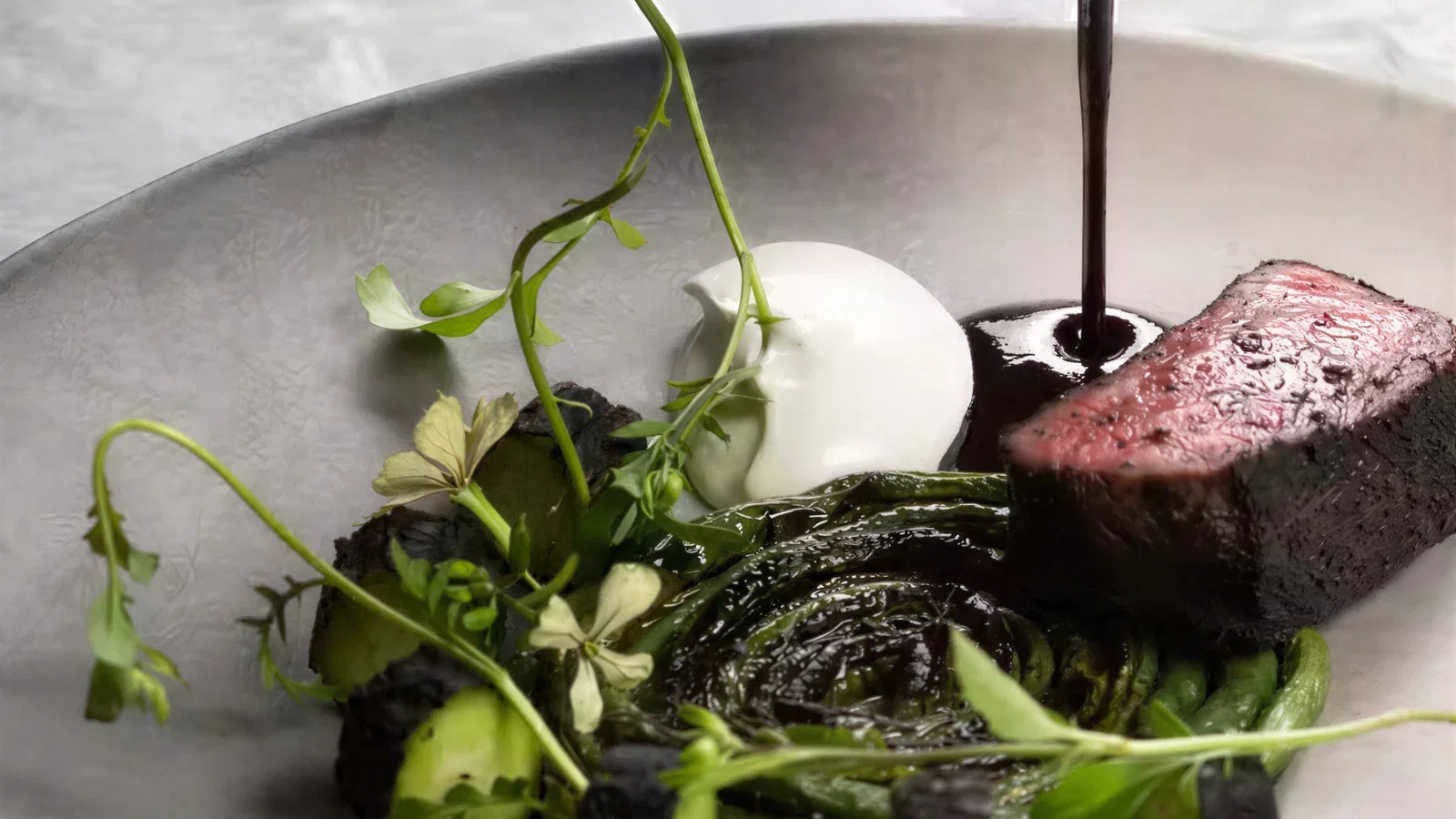 A close-up shot of a fine dining dish featuring a perfectly seared piece of meat, a dollop of white cream, and green vegetables.
