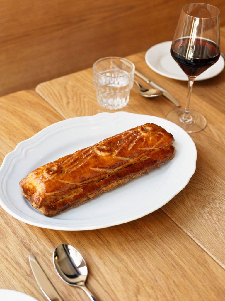 A golden-brown puff pastry on a white plate with a glass of red wine and water glass on a wooden table