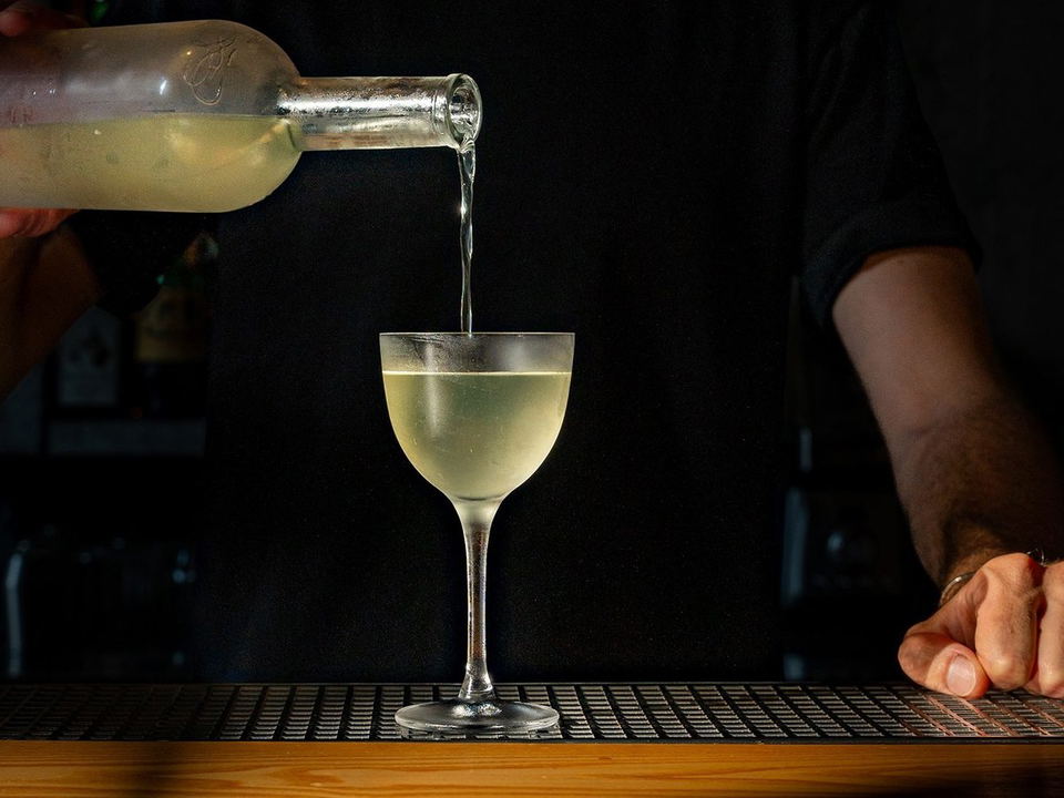 A bartender's hands pouring a pale cocktail from a bottle into a coupe glass on a bar mat