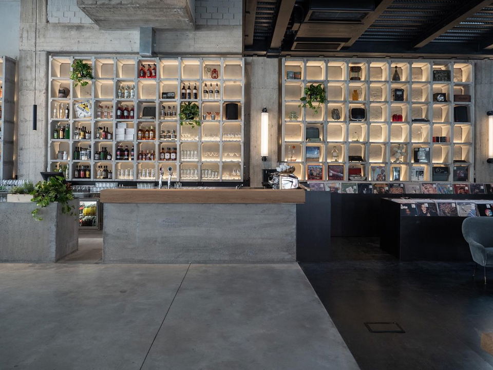 A modern industrial bar/retail space with exposed concrete, backlit cubby-hole shelving displaying bottles and products, a concrete bar counter, and p