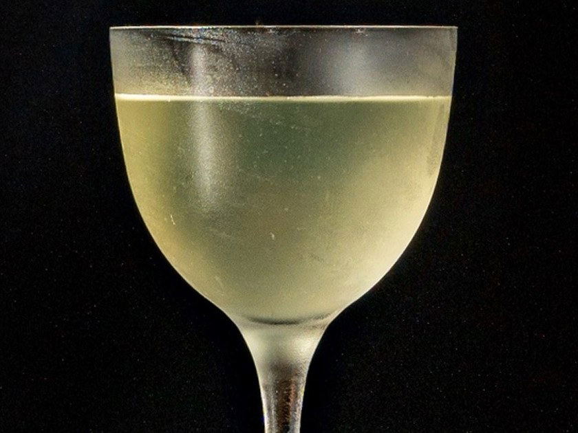 A cocktail in a coupe glass containing a pale greenish-yellow liquid against a black background