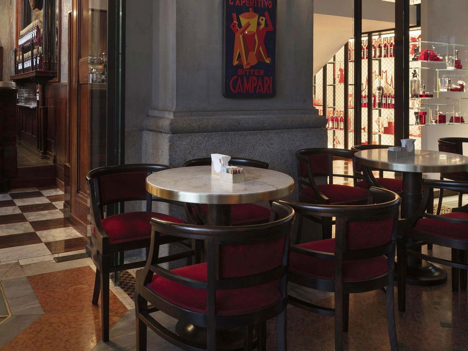 An elegant historic café interior with marble columns, ornate architectural details, vintage Campari signage, checkered flooring, and round marble-top