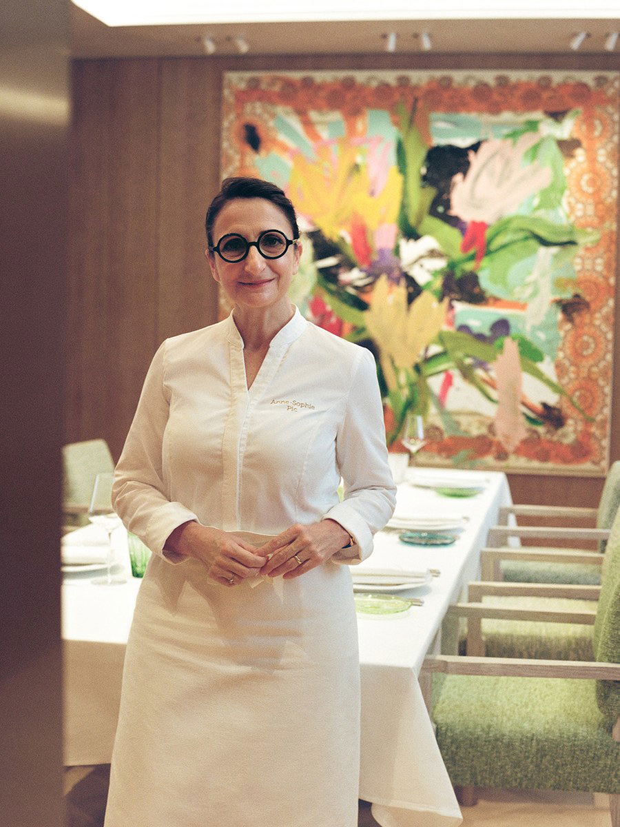 A woman in chef's whites with embroidered name stands in an upscale dining room with colorful artwork.