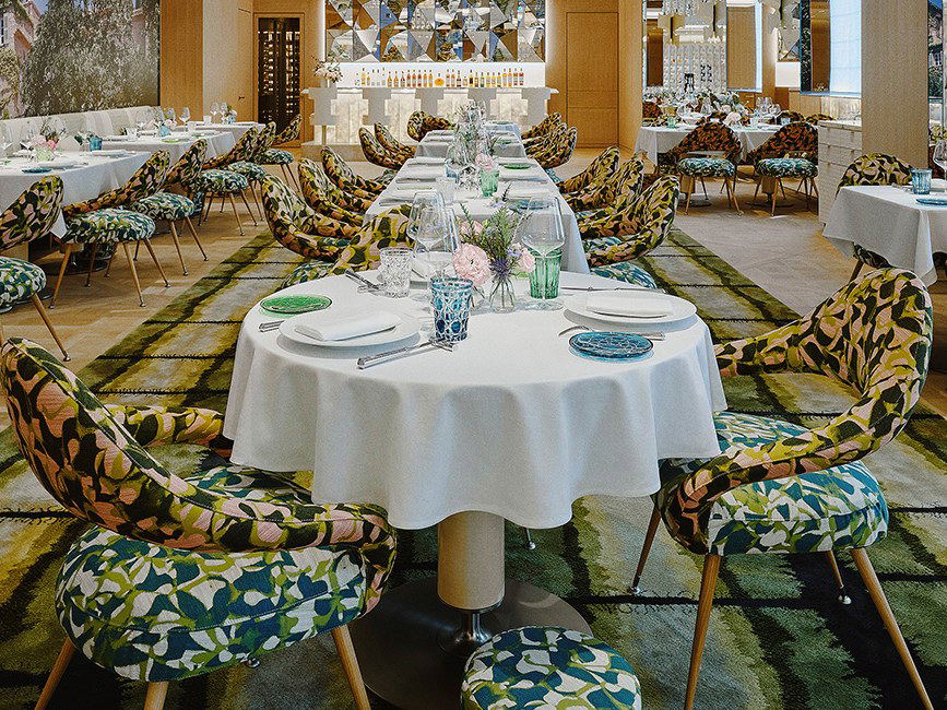 An upscale restaurant dining room with distinctive bold floral-patterned chairs in yellow/green tones, white-clothed tables with blue glassware, and m